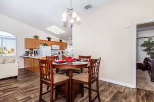 A cozy dining area featuring a round wooden table set for four with decorative plates and a centerpiece. The space is well-lit by a hanging light fixture and includes a kitchen in the background with wooden cabinets and appliances. Soft, neutral tones and a plant add warmth to the room.