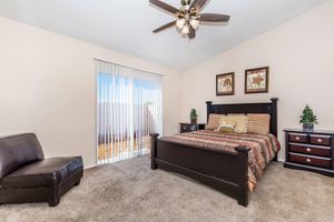 A cozy bedroom featuring a queen-sized bed with a striped comforter, wooden furniture including nightstands and a dresser, a comfortable chair, and vertical blinds allowing natural light. The walls are painted a neutral color, creating a warm and inviting atmosphere.