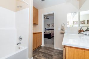 Spacious bathroom featuring a bathtub with a shower, wooden cabinets, and a white countertop. A door leads to a bedroom in the background with a ceiling fan and framed artwork on the walls. The flooring is a light brown laminate, enhancing the bright and airy atmosphere.