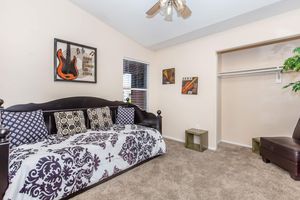 A cozy bedroom featuring a daybed with a decorative black and white bedspread, framed artwork on the walls, a small side table, and an open closet. The room has neutral-colored walls and carpet, with a ceiling fan providing airflow. Natural light streams in through a window.