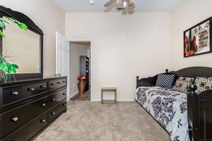 A cozy bedroom featuring a black-framed bed with patterned bedding, a black dresser, and a large mirror. There’s a small green table beside the bed, and a door leading to another room in the background. Soft carpet flooring and a guitar-themed wall art enhance the inviting atmosphere. Green plant adds a touch of nature.