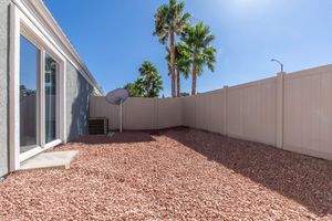A private outdoor space featuring a gravel-covered yard, surrounded by a tall fence. There are several palm trees in the background, and a satellite dish is mounted on one side. The sky is clear and blue, suggesting a sunny day. Sunlight casts shadows across the area.