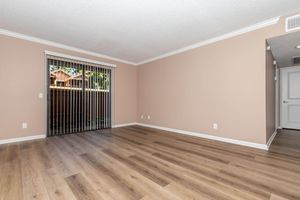 A spacious, empty room with light beige walls and wooden flooring. A large window with vertical blinds lets in natural light, revealing an outdoor view. The room has a simple, clean aesthetic with no furniture or decor, creating a blank canvas for interior design.