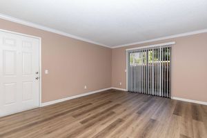 Empty room featuring light beige walls, a white door on the left, and sliding glass doors with vertical blinds on one side. The flooring is a light wood laminate. Natural light is streaming in through the sliding doors, illuminating the space.