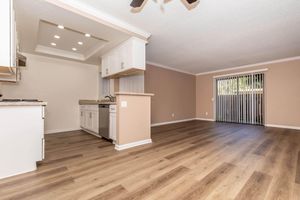 A spacious and modern living area featuring light wood flooring, a kitchen with white cabinets and a marble countertop, and a sliding glass door leading to an outdoor space. Ceiling fan and recessed lighting create a bright atmosphere in the open layout.