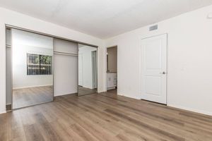 A spacious, well-lit room with light-colored walls and wooden flooring. There's a large mirrored closet along one wall, and an open door leading to another area. The room features a window with blinds, allowing natural light in. The overall aesthetic is modern and inviting.
