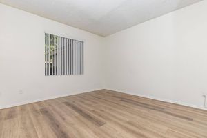 An empty room with light-colored walls and wooden flooring. A window with vertical blinds lets in natural light, and there are no furnishings or decorations in the space, giving it a clean and minimalist appearance.
