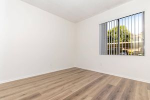 Interior view of a simple, empty room with white walls and light brown, laminate flooring. A window with vertical blinds lets in natural light, showing greenery outside. The room appears spacious, with no furniture present, creating a clean and minimalist atmosphere.