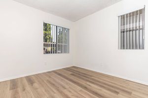 A bright, empty room with light-colored walls and laminate flooring. Two windows with vertical blinds allow natural light to enter, creating a spacious and airy feel. The room is ready to be furnished and decorated.