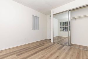 Empty room featuring light-colored walls and wooden flooring. A mirrored closet is visible on one side, along with a door leading to another area. A small window with vertical blinds is positioned on the wall, allowing natural light to enter the space. The overall atmosphere is bright and airy.