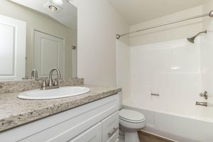 A modern bathroom featuring a white bathtub with a glass shower rod, a circular sink on a granite countertop, and a white cabinet. The walls are light-colored, and there’s a door leading to another room in the background. The overall design is clean and contemporary, emphasizing functionality and style.