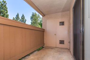 A small, covered outdoor area featuring a tan wall with a door on the right side. The door has a small window and is surrounded by concrete flooring. In the background, there are green trees visible, and the sky is clear. The scene appears to be well-lit and inviting.