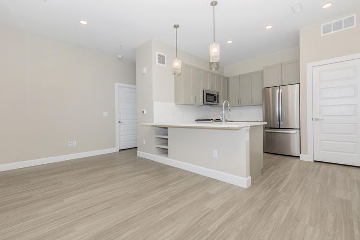 A modern kitchen and living area featuring light-colored walls, hardwood-style flooring, and a spacious layout. The kitchen is equipped with stainless steel appliances, including a microwave and refrigerator, and has contemporary cabinetry. The space includes large windows allowing natural light.