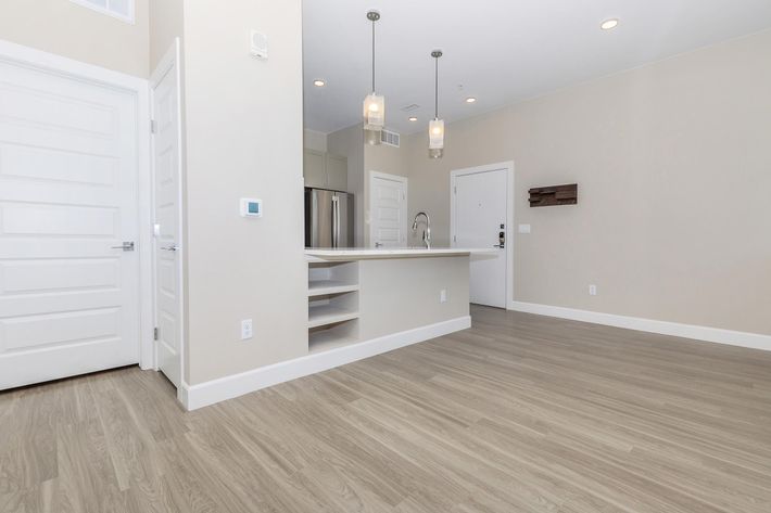 A modern kitchen area with a light-colored countertop, wooden flooring, and pendant lighting. The room features a refrigerator in the background, white cabinets, and a doorway leading to another room. Minimalist design with neutral walls and natural light.