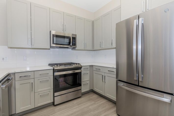 Modern kitchen featuring light gray cabinets, stainless steel appliances including a refrigerator, microwave, and oven. The spacious design includes a light-colored countertop and wood-style flooring, creating a contemporary and functional cooking space.