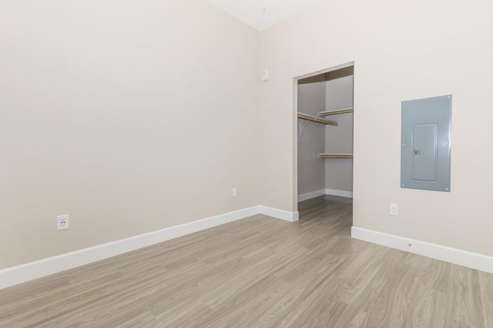 A clean, empty room with light-colored walls and a wooden floor. On the left side, there is a small closet with shelves and an open doorway. A gray electrical panel is mounted on the right wall, and there are two electrical outlets visible on the walls. The overall space has a bright and minimalist aesthetic.