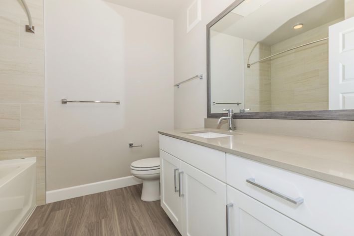 A modern bathroom featuring a light-colored vanity with a sink and mirror, a toilet, and a shower area. The walls are a soft neutral tone, complemented by light wood-like flooring. A towel bar is mounted near the sink, and the space is well-lit with natural light.
