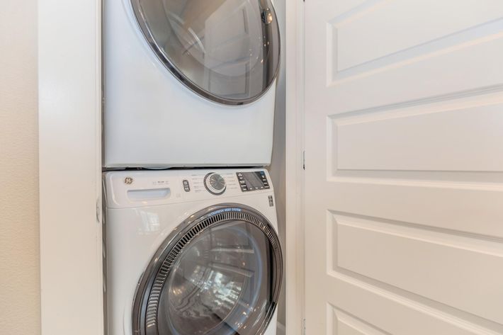 A stacked washer and dryer unit located in a narrow space beside a closed white door. The appliances are modern and feature a sleek design with control dials and a window on the dryer to view clothing inside. The wall appears painted in a light color, enhancing the brightness of the area.