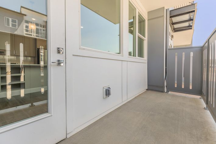 A balcony with a sliding glass door leading to an interior space. The floor is concrete, and the walls are white. On one side, there’s a gray partition with vertical slits. Reflections from the glass door reveal a modern kitchen area inside. The space is bright and airy, illuminated by natural light.