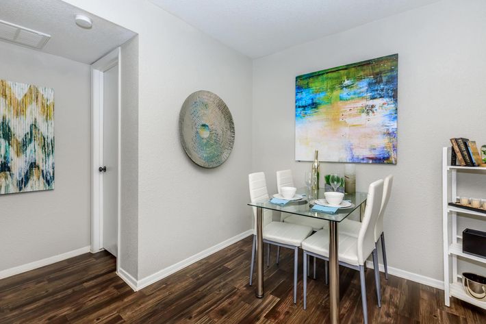 A modern dining area featuring a glass table with white chairs. On the wall is a colorful abstract painting, while another wall displays a round decorative piece. There are decorative items on a nearby white shelf, and the space has brown wooden flooring and neutral-colored walls.