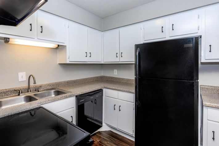 Modern kitchen with white cabinets and a black refrigerator. It features a double sink, a black dishwasher, and a black stovetop. The countertops are gray, and the flooring has a wooden appearance. The space is well-lit with overhead lighting.