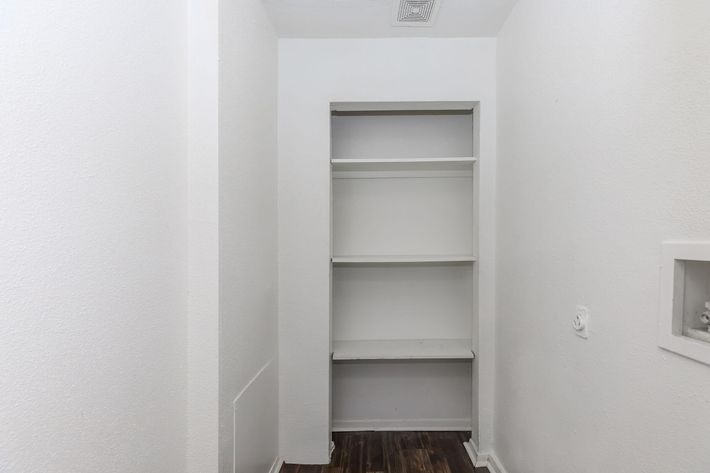 A small, empty closet with white walls and a wooden floor. There are three white shelves against one side, and a small compartment with a white frame on the opposite wall. The space is well-lit, creating a clean and minimalistic appearance.