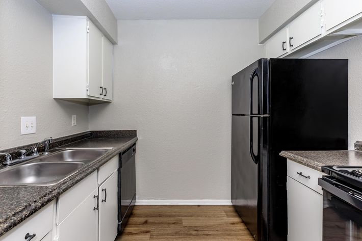 A modern kitchen featuring white cabinets, a black refrigerator, and dark countertops. The setup includes a double sink and a built-in stove, with light-colored walls and wood-like flooring, creating a clean and spacious appearance.