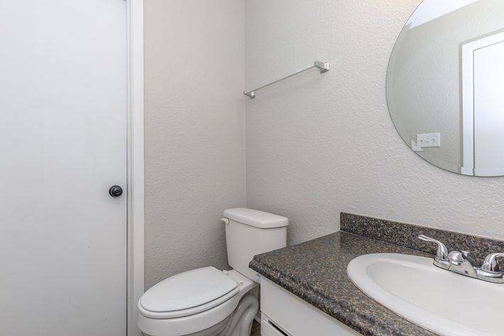 A clean bathroom featuring a round mirror above a sink with a countertop, a white toilet, and a door partially visible. The walls are painted light gray, and a towel bar is mounted on the wall. The overall setting is simple and well-maintained.