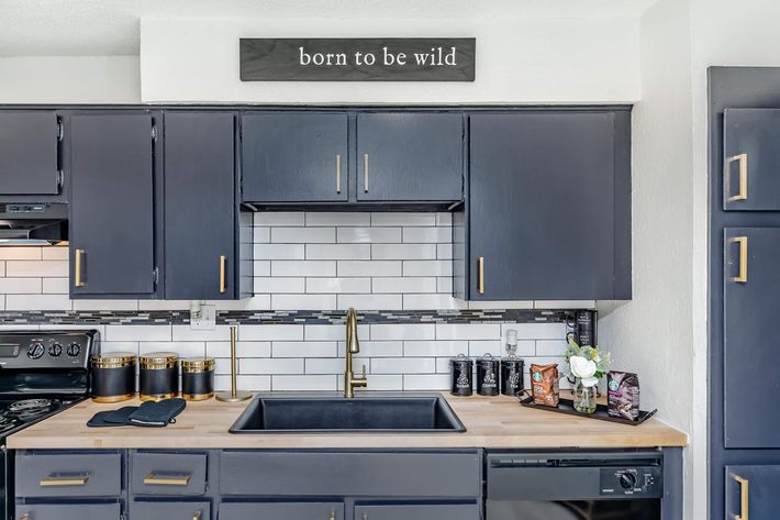 A modern kitchen featuring dark cabinetry, a black sink, and a stylish backsplash with white tiles. Above the sink is a sign that reads "born to be wild." The counter is adorned with a few decorative items, including a vase with flowers and canisters, creating a contemporary and inviting atmosphere.