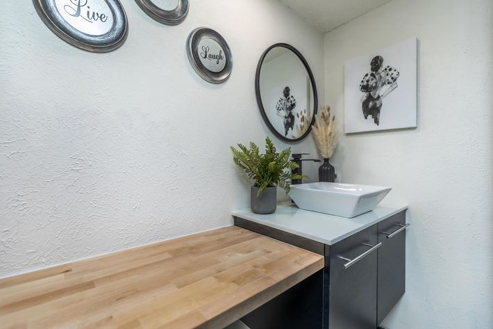 A well-designed bathroom featuring a wooden countertop, modern sink, and a stylish round mirror. The walls are decorated with black and white circular wall art and a framed picture. A potted fern and dried plants add a touch of greenery to the minimalist decor.
