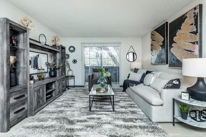 A modern living room featuring a light gray sofa, a black coffee table on a soft area rug, and a wall-mounted entertainment unit. Decorative elements include plants, vases, and large artwork. Natural light streams in through a window, enhancing the cozy ambiance.