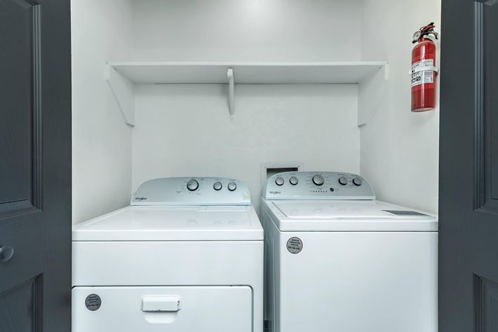 A clean laundry room featuring a white washing machine and dryer side by side. Both appliances have control panels on the front, and a fire extinguisher is mounted on the wall above. A shelf is installed above the machines for additional storage. The walls are painted in a neutral color.