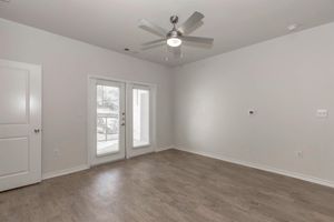 A spacious, empty room with light gray walls and wood-like flooring. It features a ceiling fan, a pair of glass doors leading to a small balcony with blinds, and a wall-mounted thermostat. The room is well-lit and has a modern, minimalist design.