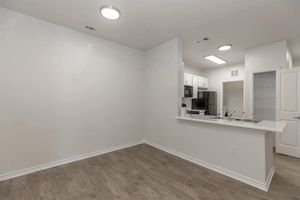 An empty, modern apartment interior featuring light-colored walls and wood-like flooring. A kitchen area with white cabinets, a black refrigerator, and a countertop is visible. Recessed lighting illuminates the space, and a door leads to another room or area.
