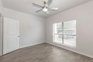 Empty room with light-colored walls and a wooden floor. A ceiling fan with five blades is mounted on the ceiling. There is a large window with blinds allowing natural light to enter, and a white door is visible on the left. The room has a clean, modern aesthetic.