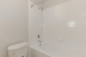 View of a clean, modern bathroom featuring a white tiled bathtub with a showerhead, a chrome faucet, and a bathtub spout. To the left, there is a white toilet. The walls are painted a light color, creating a bright and spacious atmosphere.