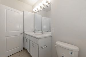 A clean, modern bathroom featuring a dual sink vanity with silver faucets, a large mirror above, white cabinetry, and a toilet. The walls are painted a light color, and the floor is tiled, contributing to a spacious and bright atmosphere. Two closed doors are visible in the background.