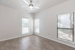 A well-lit, empty room with light-colored walls and a ceiling fan. There are two windows with blinds providing natural light, and a light wood floor. Outside the windows, greenery is visible, enhancing the room's brightness and openness. Ideal for personalizing or decorating.