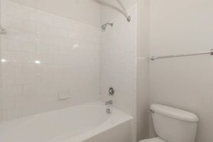 A clean bathroom featuring a white tiled shower area with a bathtub, a chrome faucet, and a wall-mounted showerhead. To the right, there is a white toilet, and the walls are painted in a light color, creating a bright and airy atmosphere.