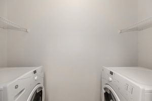 A small laundry room with two white washing machines side by side. The walls are light-colored, and there are wire shelves above the machines for storage. The space appears empty and well-lit, with a simple and clean aesthetic.