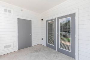 Exterior view of a small porch area featuring three doors: one solid gray door and two glass-paneled doors with gray frames. The walls are white shiplap, and there's a light fixture above the doors. The floor is concrete, and there are windows visible outside.