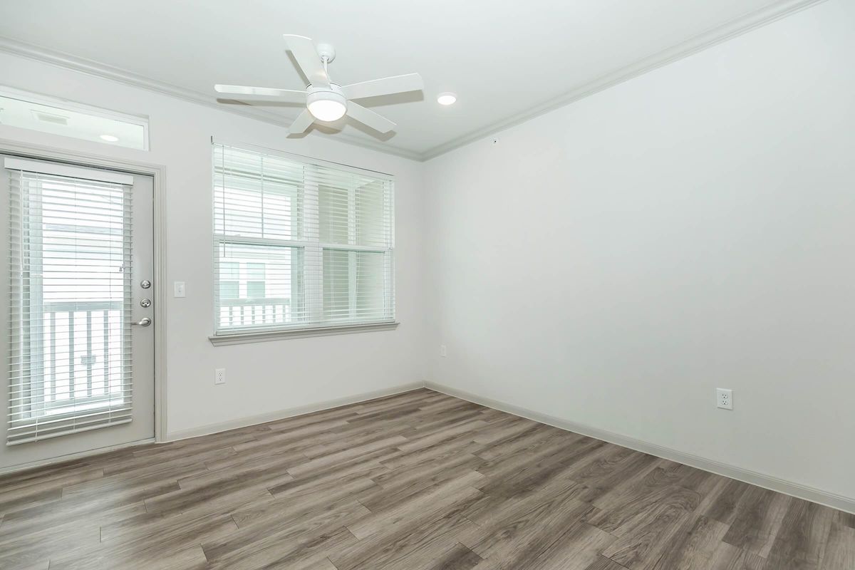 A bright, empty room featuring light-colored walls and a ceiling fan. There are large windows covered with white blinds, allowing natural light to fill the space. The flooring is a light wood finish, and there's a door leading to an outdoor area. The overall atmosphere is clean and open.