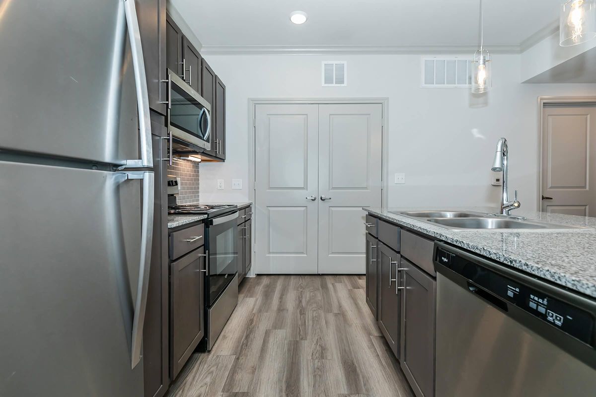 A modern kitchen featuring stainless steel appliances, including a refrigerator and oven. The granite countertops are complemented by dark cabinetry. A sink with a modern faucet is visible, and the flooring is a light wood. The walls are painted in a neutral tone, creating a contemporary aesthetic.