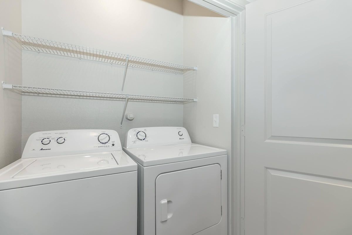 A clean laundry room featuring a stacked washer and dryer set against a light-colored wall. There are white wire shelves above the appliances for storage. A closed door is visible on the right side, contributing to a tidy and organized appearance.