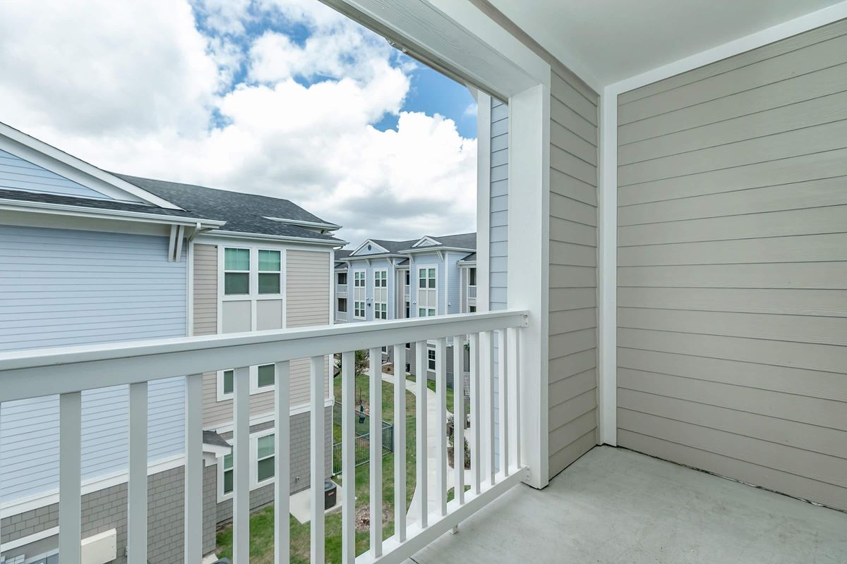 View from a balcony overlooking apartment buildings. The scene features a clear sky with scattered clouds and light gray siding on the walls. The balcony includes white railings and the ground is concrete, providing a spacious and bright outdoor area.