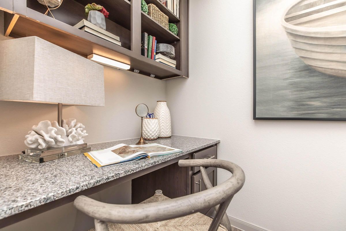 A cozy workspace featuring a granite desk with an open book, a decorative vase, and a coral accent. The shelves above hold books and plants. A stylish lamp provides soft lighting, and a framed painting of a boat hangs on the wall, creating a serene and inviting atmosphere.