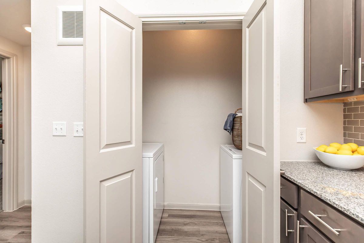 View of a laundry room through open double doors. The room features a white washer and dryer side by side against a neutral wall. A basket is placed on top of the dryer, and a bowl of lemons sits on a countertop nearby. The decor is modern with light wood flooring and contemporary cabinetry.