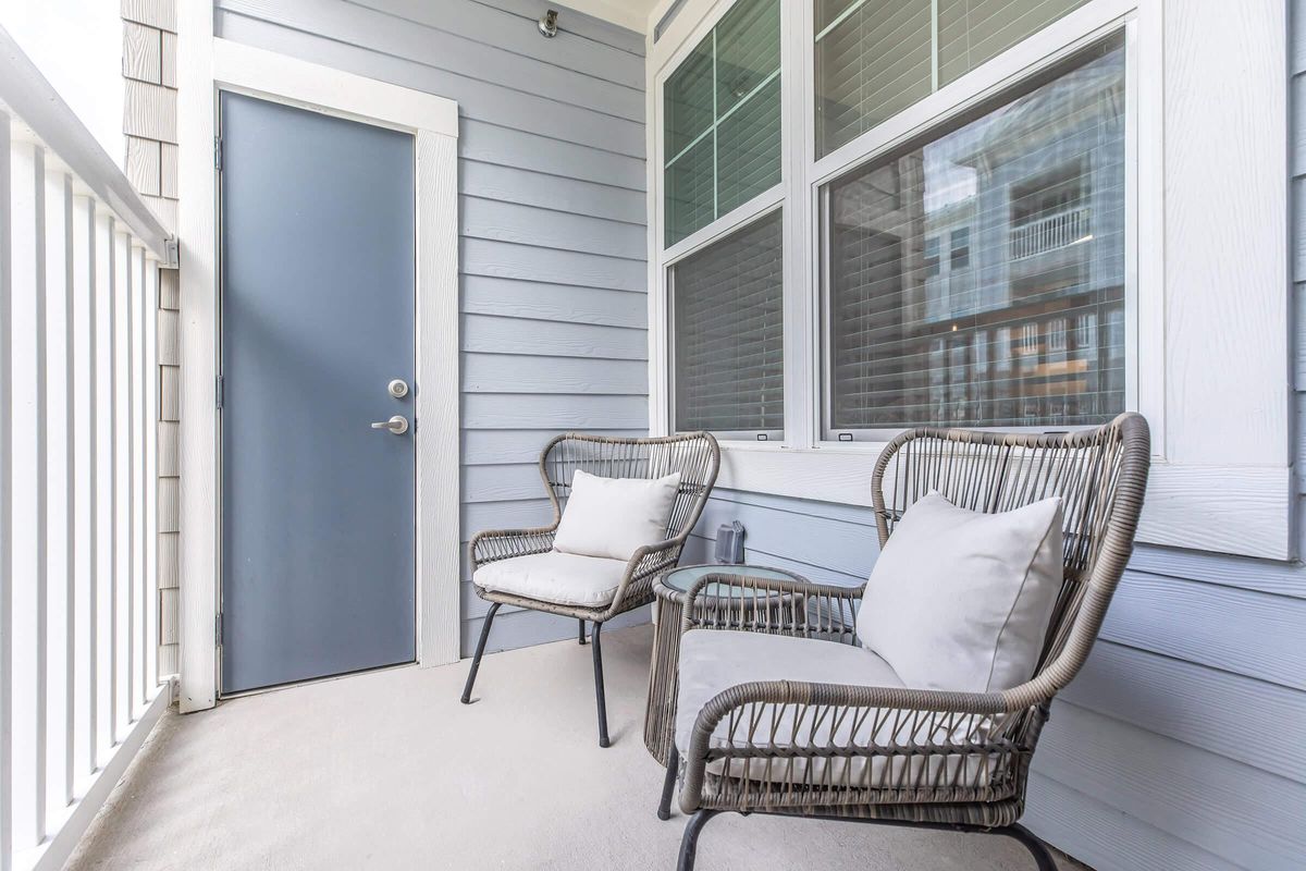 A cozy balcony area featuring two wicker chairs with cushions and a glass-top side table. The wall is painted in light blue, and there is a gray door leading inside. Large windows allow natural light to illuminate the space, creating a welcoming atmosphere.