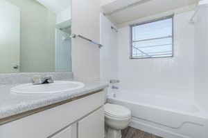 A clean and bright bathroom featuring a white bathtub and shower combination, a modern sink with a countertop, and a wall-mounted mirror. Natural light enters through a window, illuminating the neutral-colored walls and flooring.