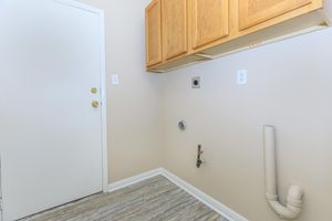 A small laundry room featuring light beige walls, a wooden cabinet above a wall-mounted appliance connection, and a door leading outside. The floor is covered in faux wood vinyl, and there is a plumbing pipe visible on the right side of the room.